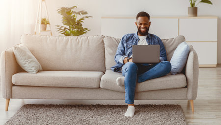 Smiling millennial black bearded guy working on laptop on sofa in living room interiorの写真素材