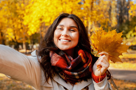 Beautiful young woman in fashionable fall clothes holding bouquet of autumn leaves and taking selfie...