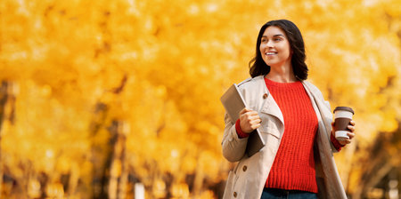 Pretty millennial woman with tablet computer and coffee to go walking at autumn parkの写真素材