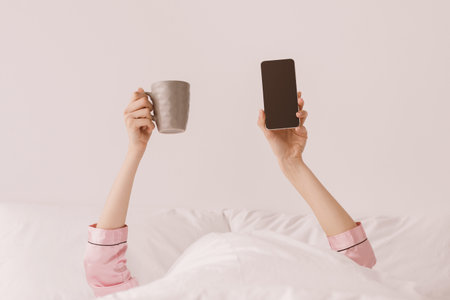 Hands of millennial caucasian lady in pink pajamas stick out from white blanket and hold cup of...