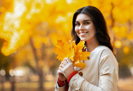 Its autumn. Portrait of joyful young woman in trendy outfit with yellow leaves looking at camera in...