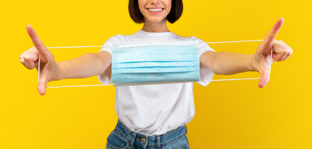 After covid. Happy woman holding medical face mask in hands and smiling at camera over yellow...