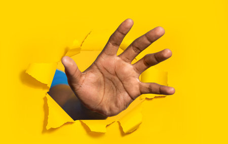 African american man reaching through hole in torn yellow paper, trying to grab something, closeupの写真素材