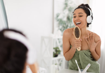Happy young woman in headphones enjoying music, singing and dancing, using hairbrush as mic, having fun near mirrorの写真素材
