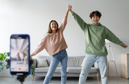 Happy young Asian couple making video on cellphone, dancing in front of camera at home