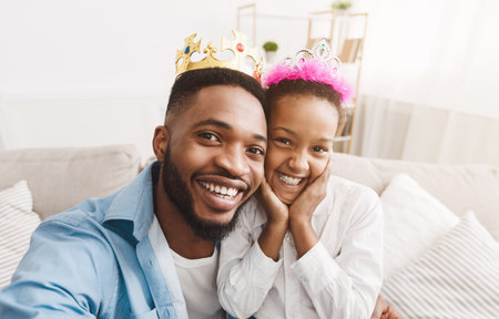 Joyful afro father and daughter wearing crowns and taking selfieの写真素材