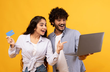 Joyful indian couple with laptop and credit card emotionally reacting to online profit, posing over...
