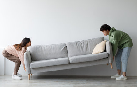 Side view of cheery young Asian couple moving sofa at home, replacing furniture in living room, blank spaceの写真素材