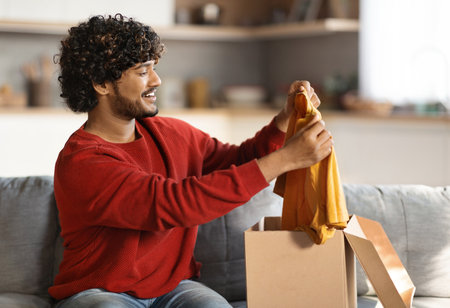 Happy Young Indian Guy Unboxing Parcel With New Clothes At Home