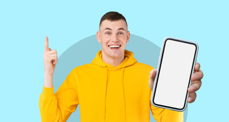 Smiling Young Man in Yellow Hoodie Holding Smartphone in Front of Light Blue Background