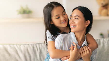 Happy asian little girl hugging her beautiful mother and smiling to each other, sitting on sofa in living roomの写真素材