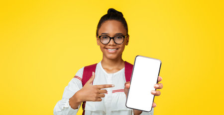 Satisfied smart adolescent black lady pupil in glasses with backpack shows finger at smartphone with blank screenの写真素材