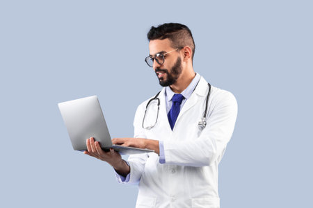 Arabic Male Doctor Using Laptop Standing Over Blue Background, Vertical