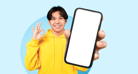 Young Man in Yellow Hoodie Happily Shows Smartphone With Blank Screen, Against Blue Background at...