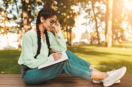 Happy indian female student sketching while sitting on bench in park outdoors, free space. Studentship conceptの写真素材