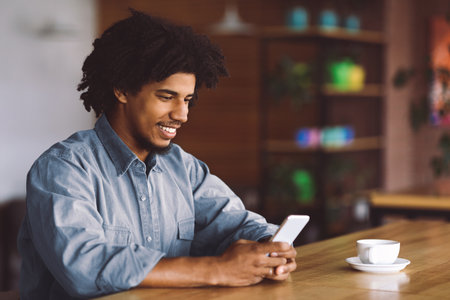 Young African American Guy Using Smartphone While Sitting At Tablet In Cafeの写真素材