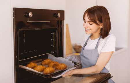 Young housewife taking out tray with fresh baked croissants from oven in kitchen, enjoying cooking homemade pastryの写真素材