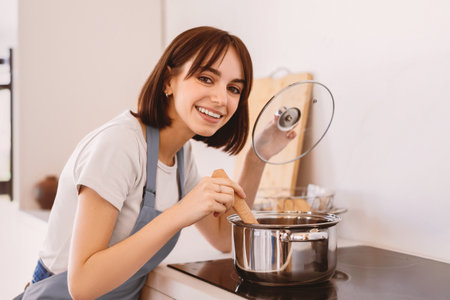 Happy lady tasting soup while cooking dinner in a pot, standing in modern kitchen and smiling at camera, wearing apronの写真素材