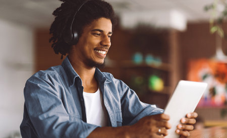 Happy Black Guy Wearing Wireless Headphones Using Digital Tablet At Cafe