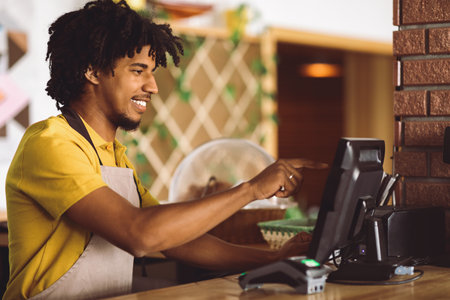 Smiling curly attractive young black guy bartender in apron behind bar counter with terminal works...