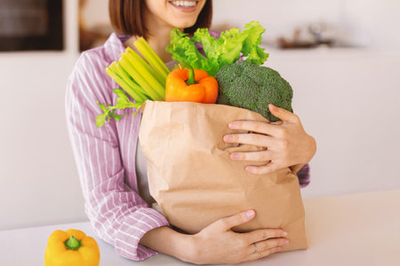 Young woman holding grocery shopping bag full of organic vegetables, standing in kitchen interior,...