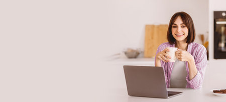 Portrait of happy lady using laptop and holding cup, drinking hot coffee in the morning, sitting in modern kitchenの写真素材