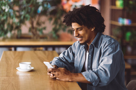 Glad happy curly handsome millennial black man typing at smartphone at table in modern cafe interiorの写真素材