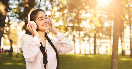 Happy indian student girl resting in campus with laptop, wearing headphones and listening music, sitting on benchの写真素材
