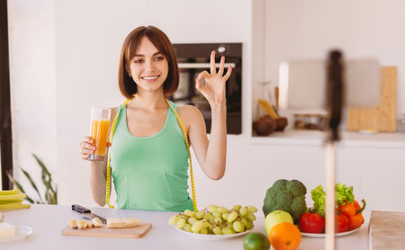 Happy fit woman drinking fresh detox and talking at smartphone camera, showing ok gesture, shooting video for vlogの写真素材