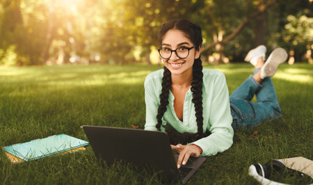 Happy indian female student studying outdoors, lying with laptop computer on lawn, smiling at...