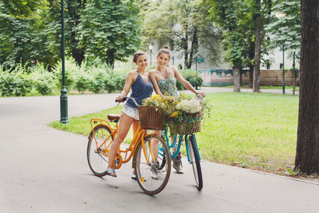 Happy boho chic girls ride together on bicycles in parkの写真素材