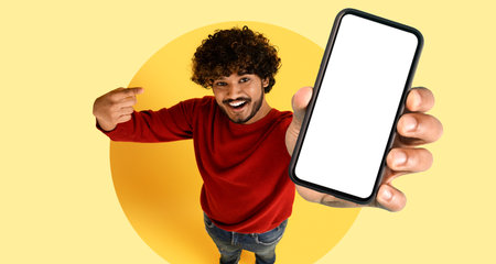 Young Man Happily Showcasing a Blank Smartphone Screen Against a Bright Yellow Background in a...