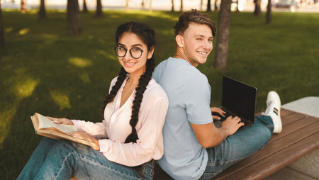 Modern students using laptop and book, learning outdoors, sitting back to back on bench in park outsideの写真素材