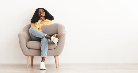 Pretty black lady sitting in armchair with laptop, copy spaceの写真素材
