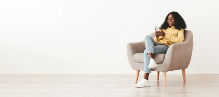Relaxed pretty black woman sitting in arm chair with cellphoneの写真素材