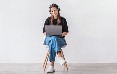 Cheery Caucasian woman in headphones having online video call on laptop, sitting on chair against white wallの写真素材