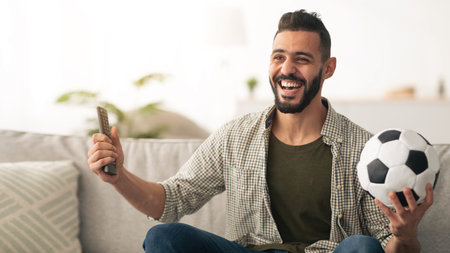 Happy Man Celebrating While Watching Soccer at Home With a Ball and Remote Controlの写真素材