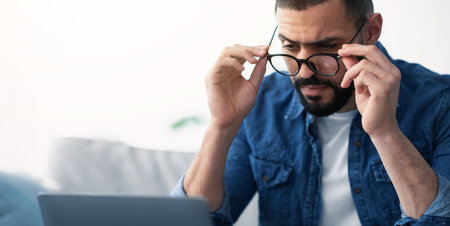 Man Adjusts Glasses While Focusing on Laptop Screen in a Cozy Indoor Settingの写真素材