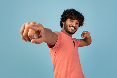 Attractive Young Indian Man Pointing at Camera and Smiling on Blue Backdrop