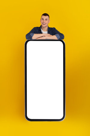 Man Smiling and Leaning on a Large Blank Smartphone Against a Bright Yellow Backgroundの写真素材
