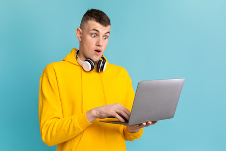 Young Man in Yellow Hoodie Surprised While Using Laptop With Headphones on a Blue Background
