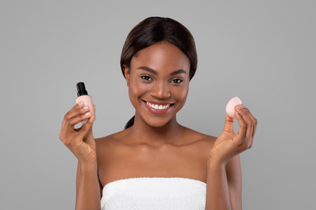 Woman Smiling While Holding a Cosmetic Foundation Bottle and Makeup Sponge in a Neutral Settingの写真素材