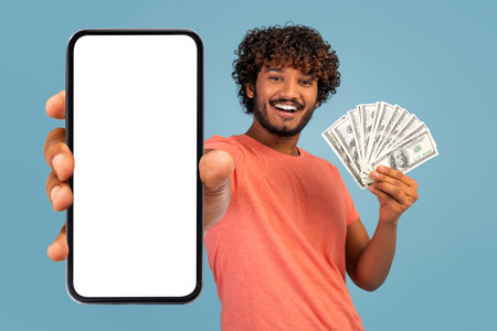 Wealthy Young Indian Man Displaying Smartphone and Cash in Bright Studio Settingの写真素材