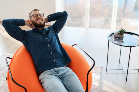 Man Relaxing in a Modern Orange Chair While Enjoying a Peaceful Moment at Home in Natural Lightの写真素材