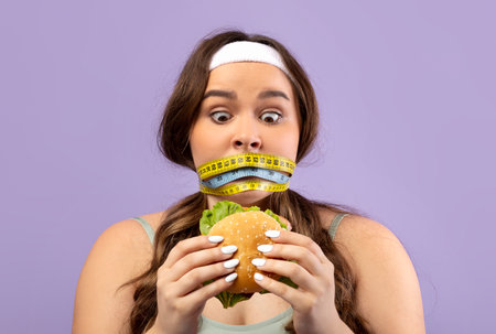 Millennial Woman in Sports Uniform Shocked by Burger While Measuring Waist With Tapeの写真素材
