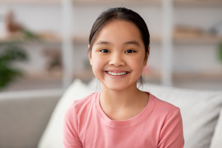 Smiling Girl With Long Black Hair in a Pink Shirt Sitting on a Couch in a Bright, Cozy Roomの写真素材