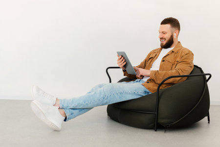 Man Enjoying Leisure Time on a Soft Chair While Browsing on a Tablet in a Modern, Minimalistic Indoor Spaceの写真素材
