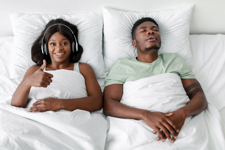 Couple Relaxing in Bed, One Enjoying Music While the Other Sleeps Peacefully in a Cozy Bedroomの写真素材