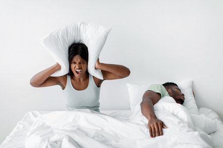 Frustrated Woman Covering Ears With Pillow While Partner Sleeps in a Messy Bed at Homeの写真素材
