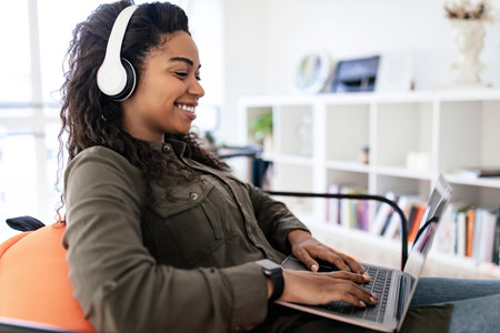 Woman Wearing Headphones Works on Laptop While Sitting in Modern Living Room With Bookshelves and...